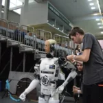 Technicians preparing a humanoid robot during a robotics event inside an indoor arena, with seating stands and equipment visible in the background.