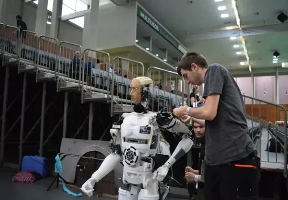 Technicians preparing a humanoid robot during a robotics event inside an indoor arena, with seating stands and equipment visible in the background.