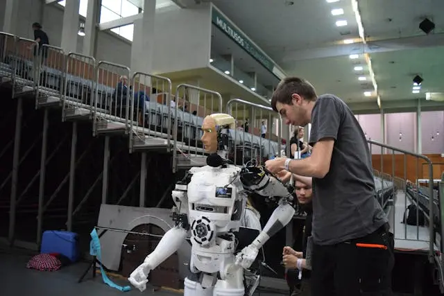Technicians preparing a humanoid robot during a robotics event inside an indoor arena, with seating stands and equipment visible in the background.